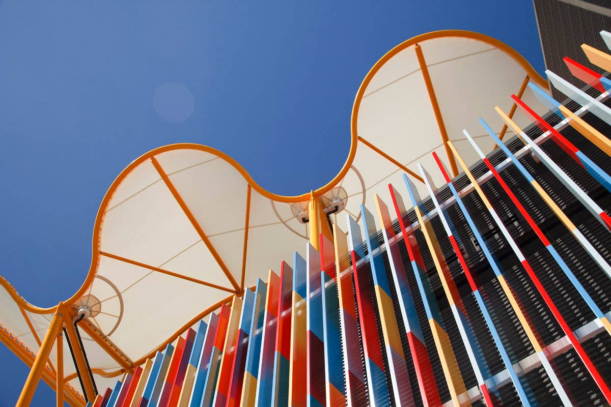 Upward view of People First Stadium roof canopy with colourful vertical façade panels against a clear blue sky.