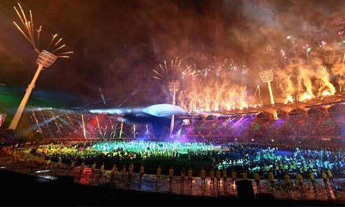 Nighttime stadium event with fireworks lighting up the sky above the field, as a large crowd gathers around a central stage under floodlights.