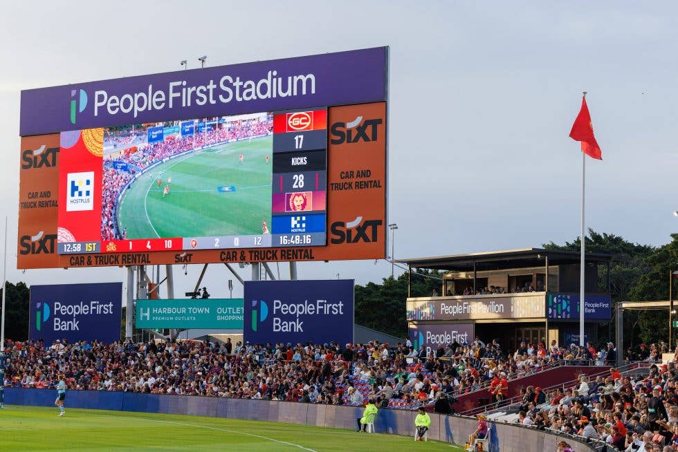 Large digital scoreboard at People First Stadium displaying live match footage and scores above a packed grandstand, with the People First Pavilion visible to the right.