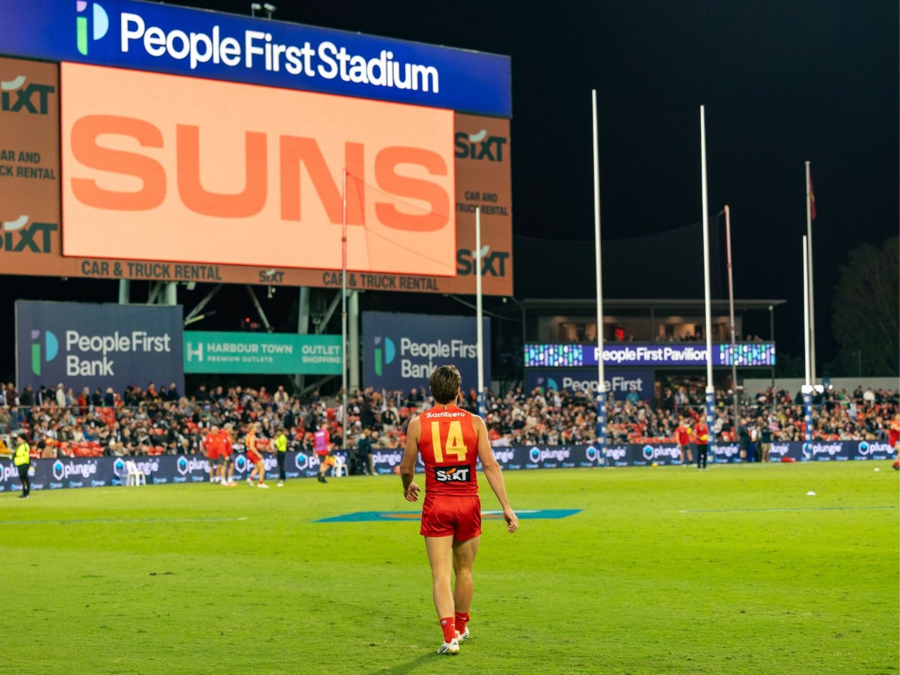 Gold Coast Suns player wearing number 14 walks across the field at People First Stadium during a night match, with the stadium scoreboard displaying “Suns” and spectators in the background.