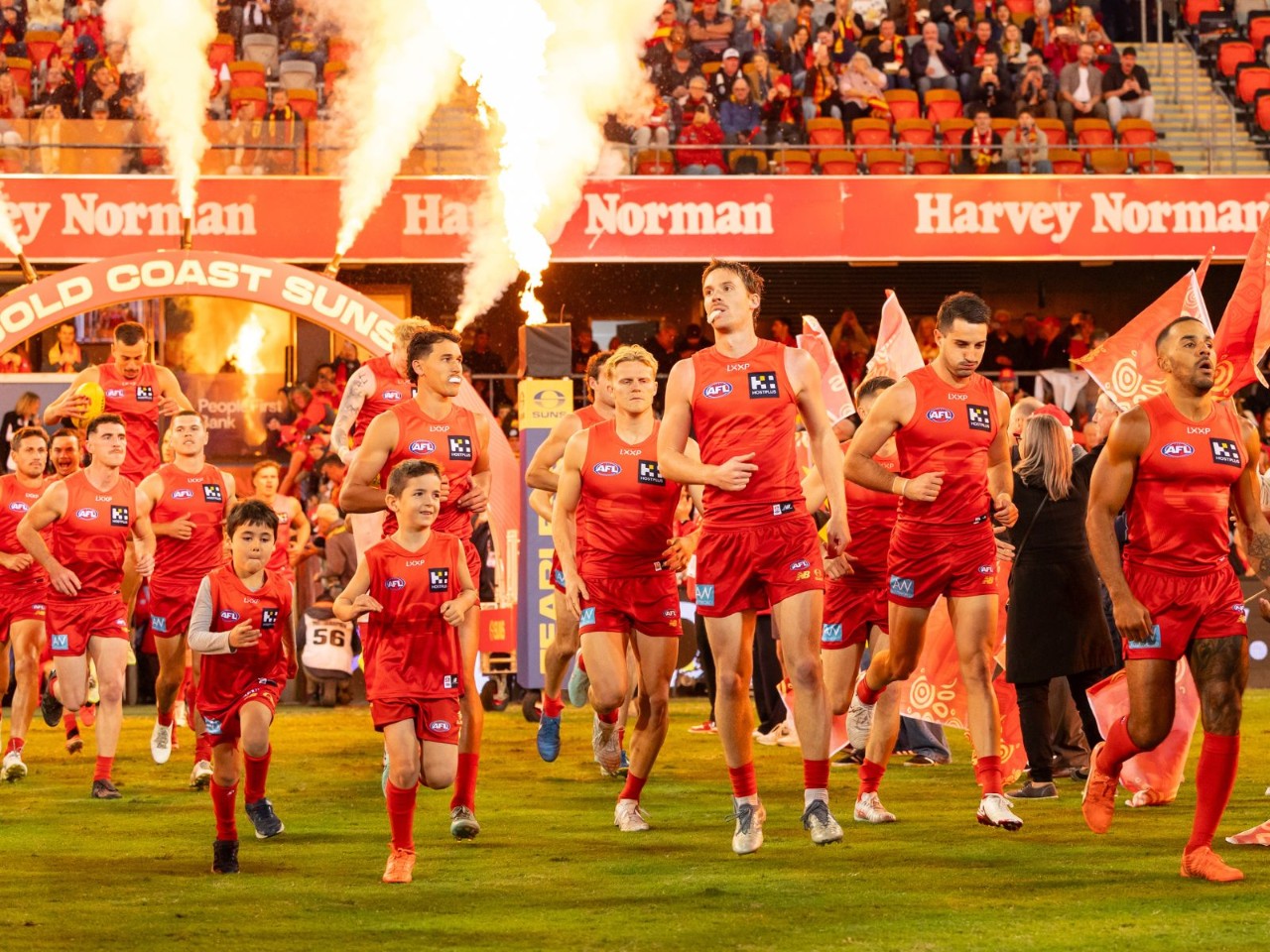  Gold Coast Suns players run onto the field with junior mascots at People First Stadium during a night match, with crowd and stadium signage visible in the background.