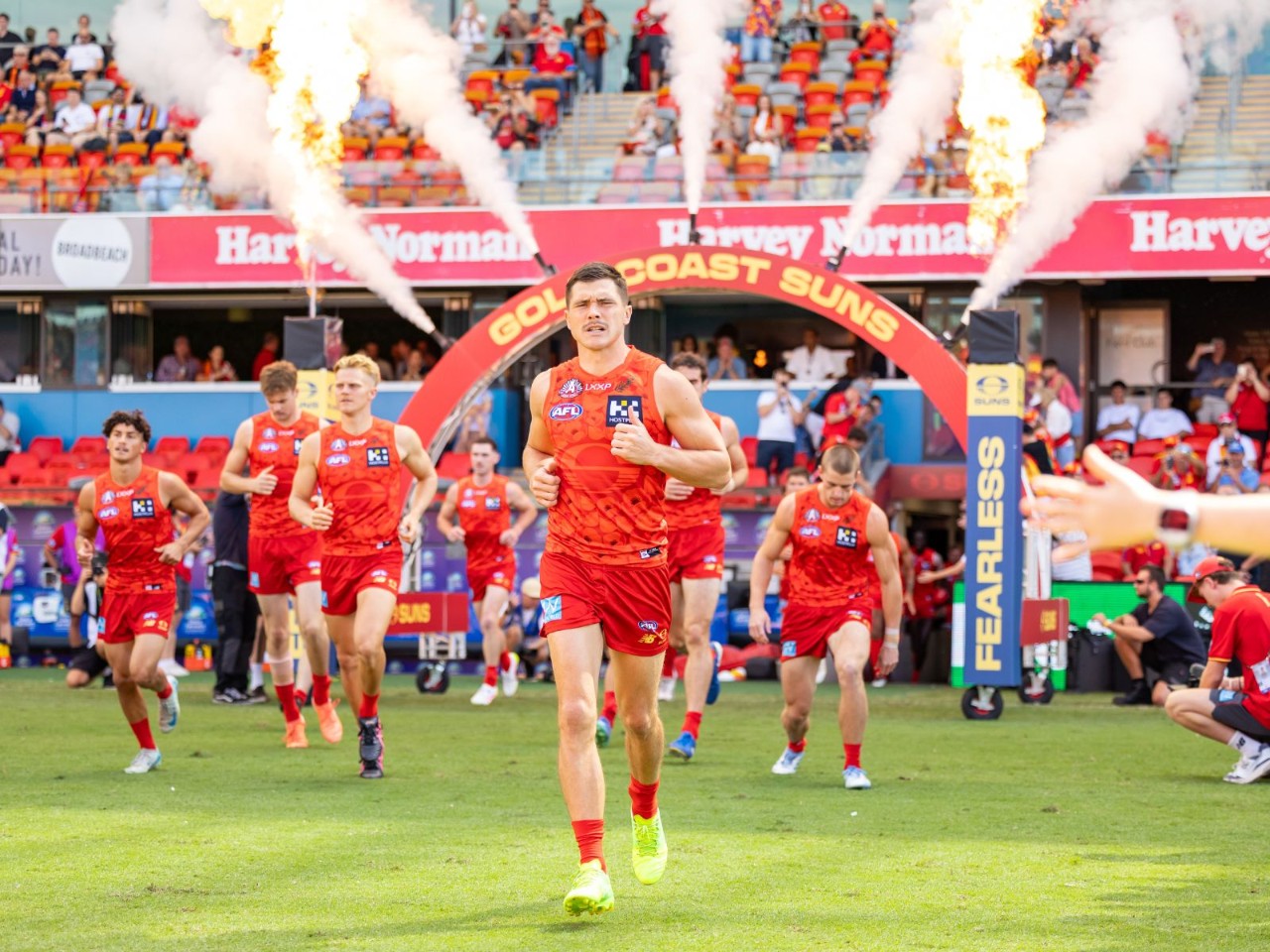 Gold Coast Suns players run onto the field through a branded arch at People First Stadium, with smoke effects and spectators visible in the background.