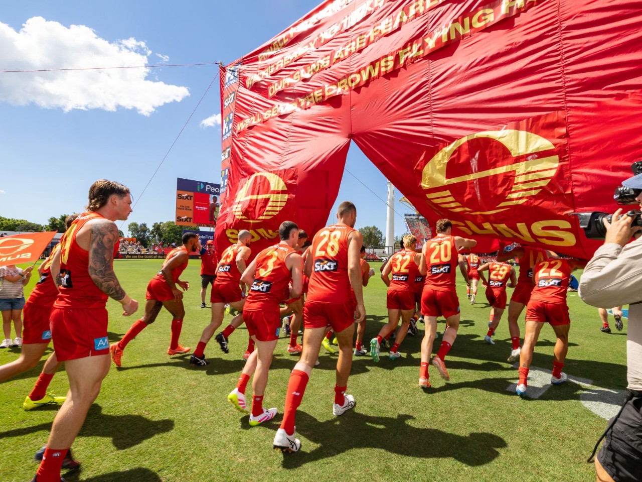 Gold Coast Suns players run through a large red Suns banner onto the field at People First Stadium before a match, with clear blue sky overhead.