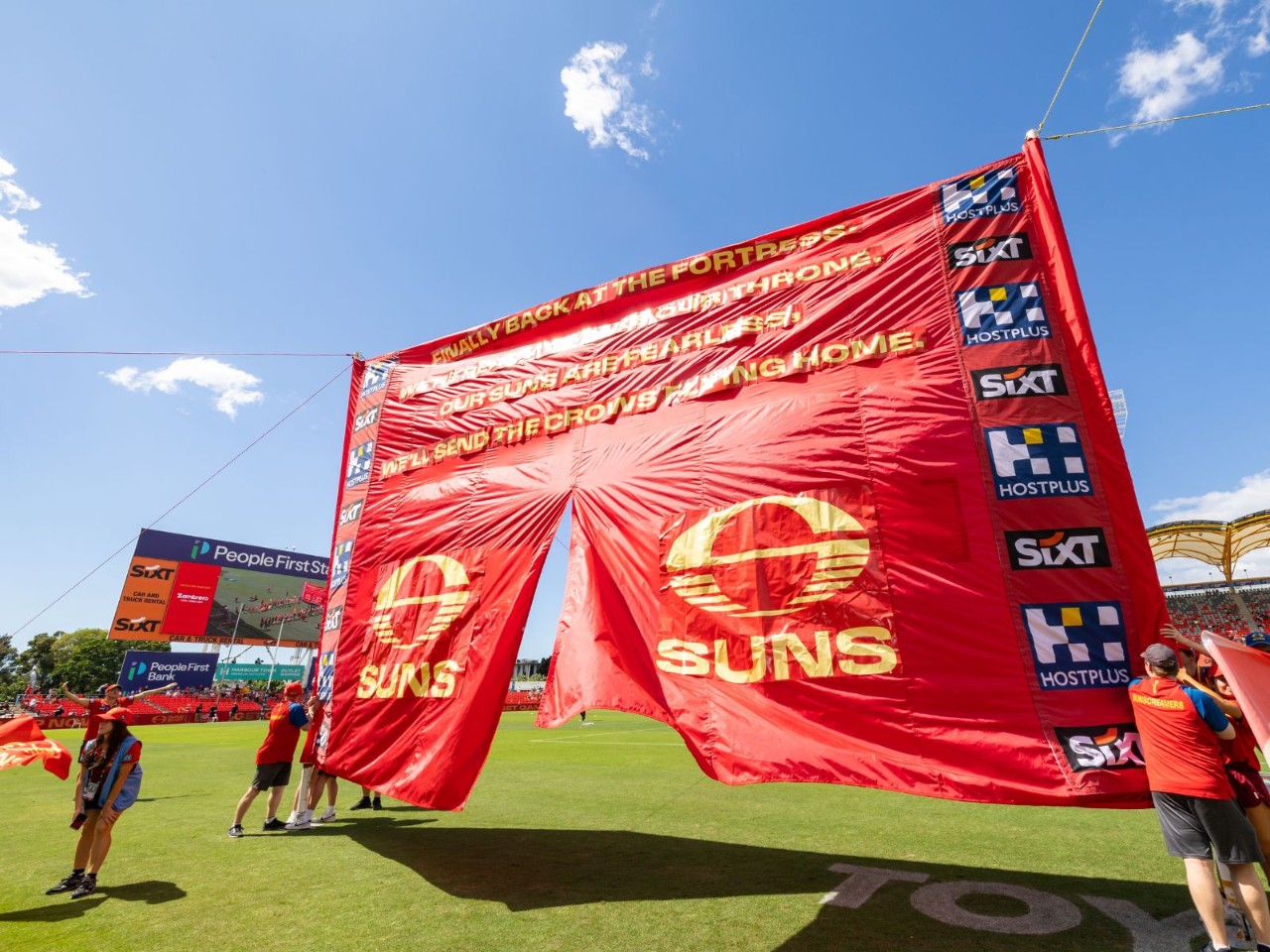 Gold Coast Suns players run through a large red Suns banner on the field at People First Stadium under a bright blue sky, with sponsor logos displayed on the banner.