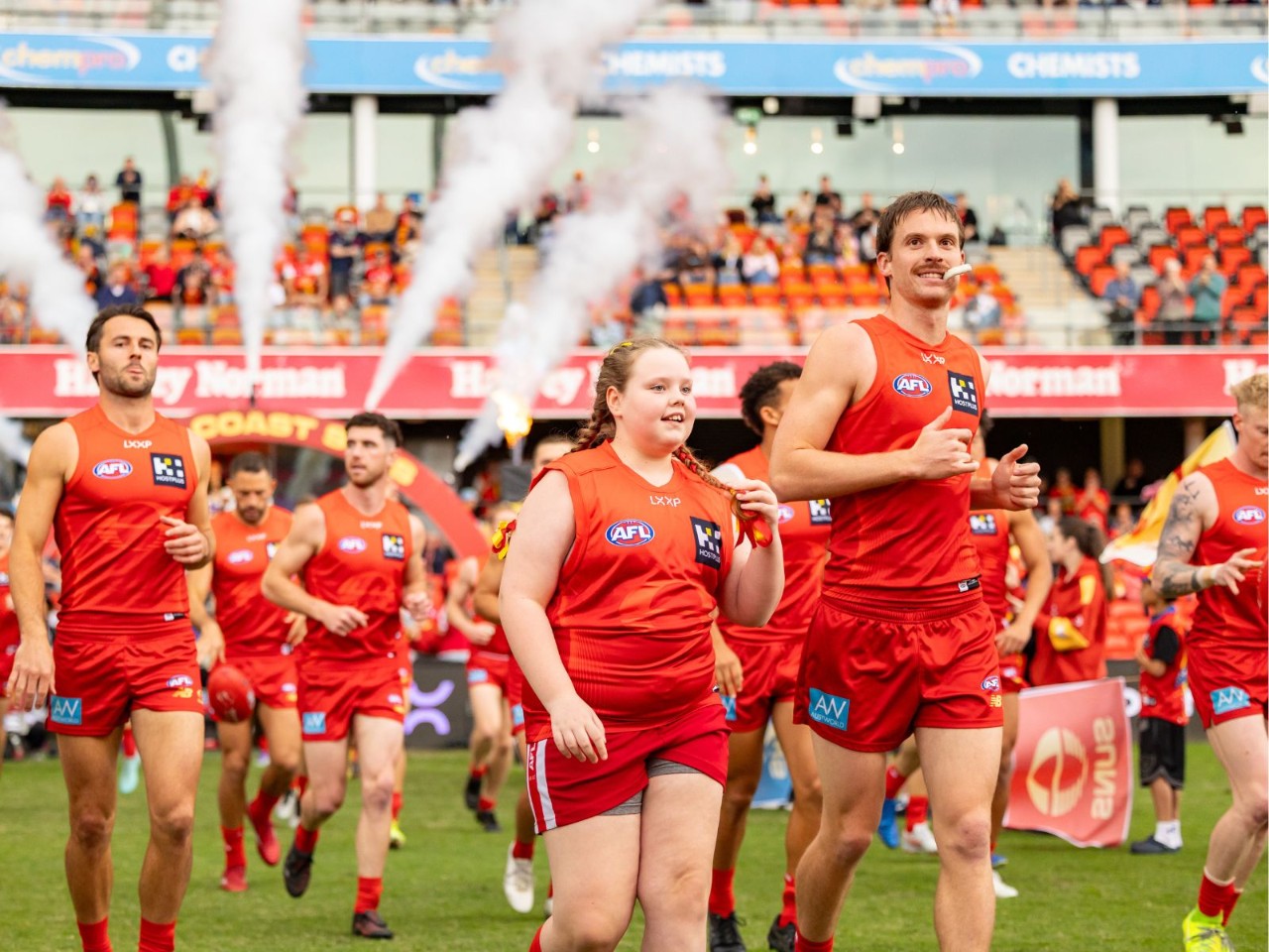 Gold Coast Suns players in red uniforms run onto the field at People First Stadium alongside a junior participant, with spectators visible in the stands behind them.